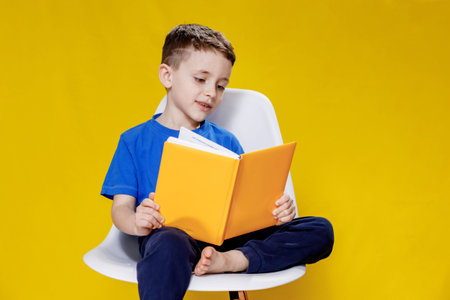 Little cheerful blond green-eyed boy 5-6 years old in a stylish blue T-shirt holding book and reading on yellow wall background, children's studio portrait.の写真素材