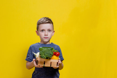 The boy is holding a basket with fresh vegetables on a yellow background. Basket with broccoli, cauliflower, cherry tomatoes, cucumber, pepper. Vegan and healthy conceptsの写真素材