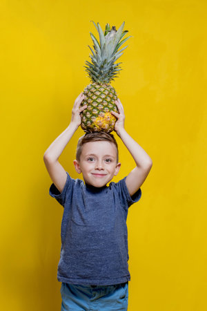 Child boy holding pineapple while posing on yellow background with copy space. Concept of summer and healthy vegan food.の写真素材