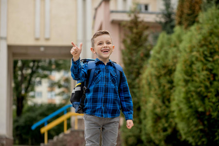 Cheerful mischievous Caucasian schoolboy in blue shirt and school jacket posing showing approval gesture, class. back to schoolの写真素材