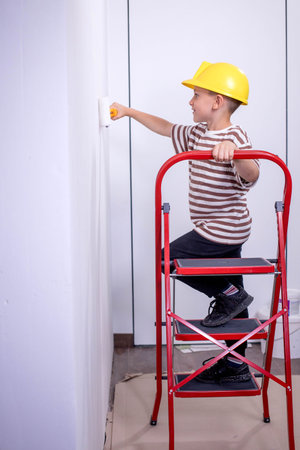 A child builder stands on a ladder and paints white walls in an apartment. A child with a paint roller during renovation.の写真素材
