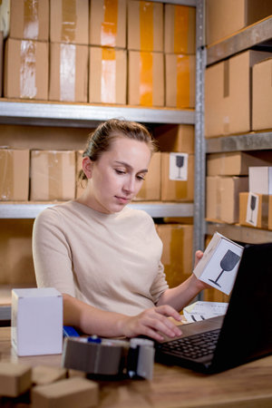 Portrait of woman entrepreneur holding smartphone and typing something at the keyboard while preparing information about parcel. Post service and small business conceptの写真素材