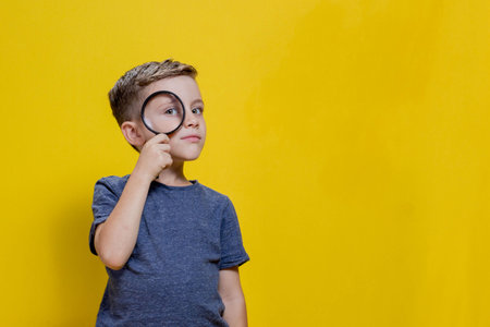 Positive curious schoolboy in casual clothes looking at camera through magnifying glass while standing on yellow background. Copy space.の写真素材