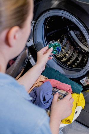 Woman putting laundry detergent capsule into washing machine indoors, closeup. Washing clothes.The...
