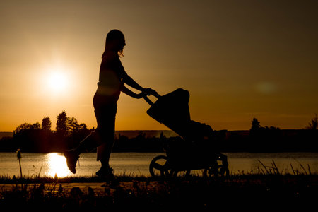 Silhouette of a mother pulling a baby carriage against the sunset near the lake.の写真素材