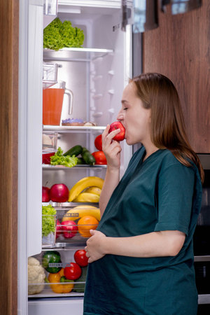 Pregnant woman is eating an apple near the refrigerator.の写真素材