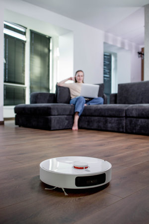 Robotic vacuum cleaner cleaning the room while woman working with laptop computer on sofa.の写真素材