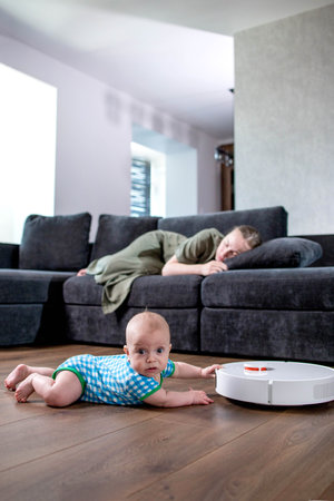 Cute young woman is sleeping on sofa in living room while little child is playing with robot vacuum cleaner on floor at homの写真素材