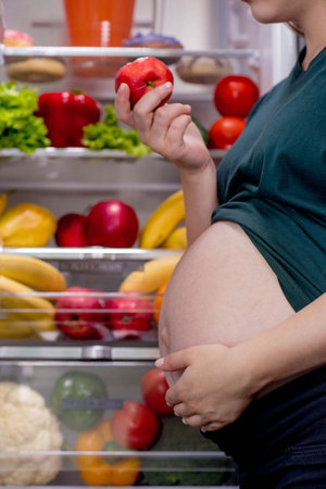Pregnant woman is eating an apple near the refrigerator.の写真素材