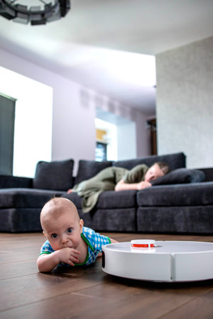Cute young woman is sleeping on sofa in living room while little child is playing with robot vacuum cleaner on floor at homの写真素材