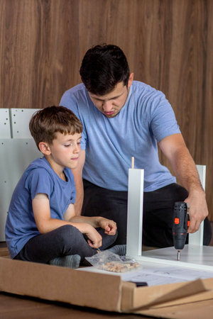 The son helps dad to collect furniture, there are tools and boards nearby. Assembling furniture at...
