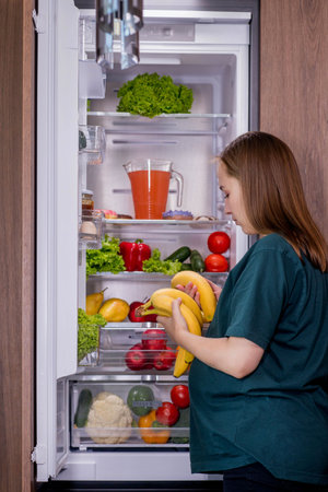 Hungry pregnant woman takes a banana from the refrigerator.の写真素材