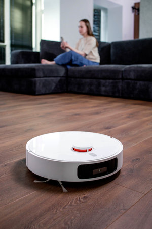 A robot vacuum cleaner cleans a room while a woman looks at her phone.の写真素材