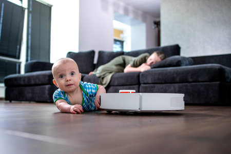Mother is sleeping on sofa in living room while little child is playing with robot vacuum cleaner on floor at home.の写真素材