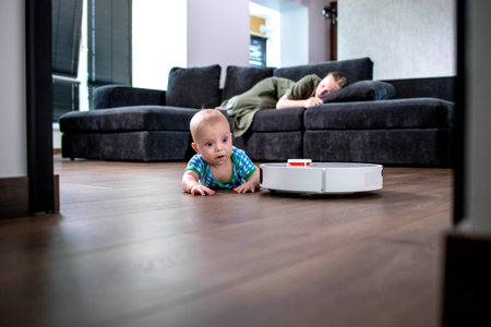 Mother is sleeping on sofa in living room while little child is playing with robot vacuum cleaner on floor at homeの写真素材