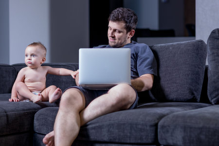 Dad sitting with his baby boy on a sofa and working on a laptopの写真素材