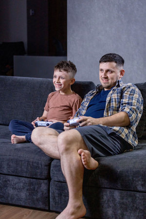 Father and his little son sit on sofa in living room playing video games together at homeの写真素材