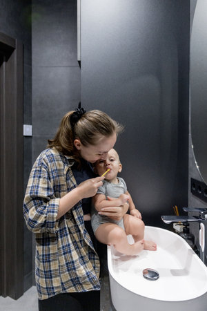 A woman is diligently brushing her teeth while simultaneously holding a baby in her arms in a bathroom space that is clean and tidyの写真素材