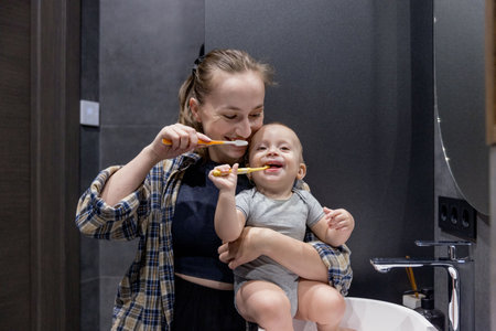 A woman is diligently brushing her teeth while simultaneously holding a baby in her arms in a bathroom space that is clean and tidyの写真素材