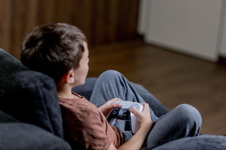 Boy is sitting on sofa at home and playing video games with his joypad