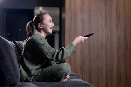 A woman comfortably positioned on a cozy couch is actively holding a remote control in her hand, ready to change the channel or volumeの写真素材