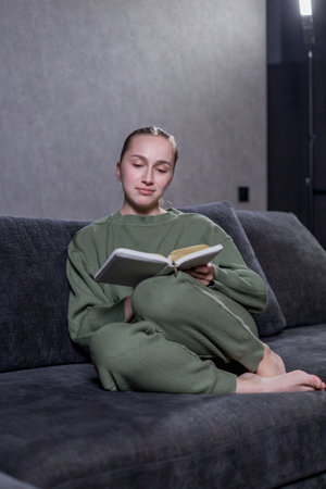 A woman is comfortably sitting on a cozy couch, fully absorbed in reading an interesting book on a pleasant afternoonの写真素材