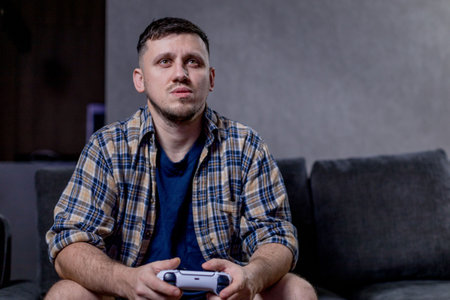 A man is comfortably seated on a couch while holding a video game controller in his hands, absorbed in the game he is playingの写真素材