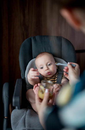Mother feeding her little baby in kitchen at home. Baby's first food.の写真素材