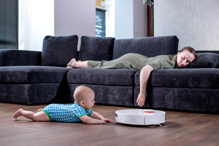 Mother is sleeping on sofa in living room while little child is playing with robot vacuum cleaner on floor at home.の写真素材