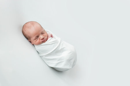 A precious newborn baby is gently swaddled in a soft, white blanket, providing warmth and comfort to the little one while they sleep peacefullyの写真素材