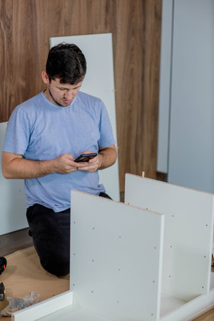 A young man kneels while intently looking at the furniture assembly instructions in his phone. Assembling furniture at home.の写真素材