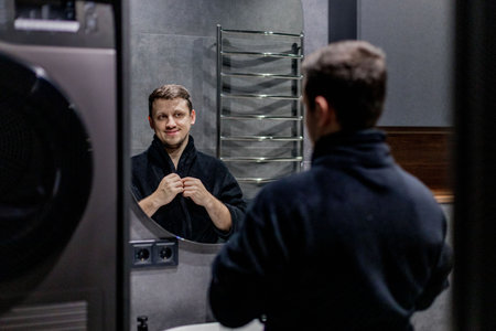 A man dressed in a comfortable bathrobe is standing and gazing at his reflection in a large bathroom mirror, assessing his appearanceの写真素材