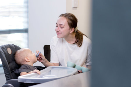 A woman is carefully feeding a small baby who is sitting in a high chair using a spoon to deliver the food to the toddlerの写真素材