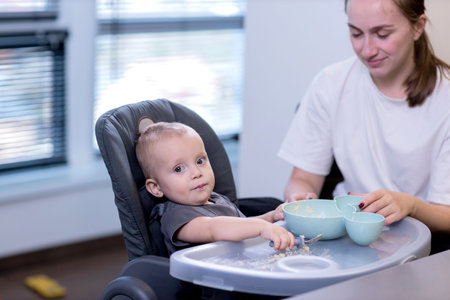 A woman is carefully feeding a small baby who is sitting in a high chair using a spoon to deliver the food to the toddlerの写真素材