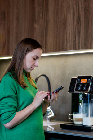 Young woman looking at phone in kitchen and making coffee in coffee makerの写真素材