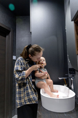 A woman is diligently brushing her teeth while simultaneously holding a baby in her arms in a bathroom space that is clean and tidyの写真素材