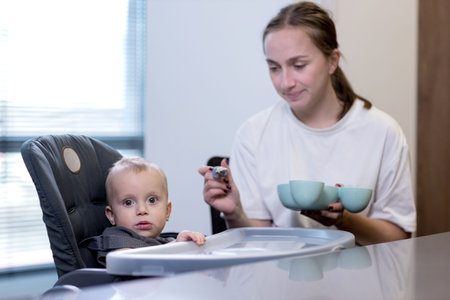 A woman is carefully feeding a small baby who is sitting in a high chair using a spoon to deliver the food to the toddlerの写真素材