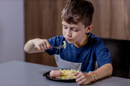 Little boy is happily eating pasta farfalle with sausage in the kitchen.の写真素材