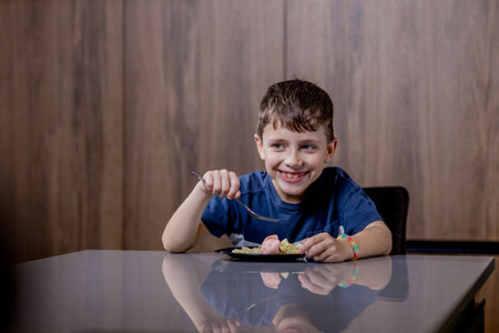 Little boy is happily eating pasta farfalle with sausage in the kitchen.の写真素材