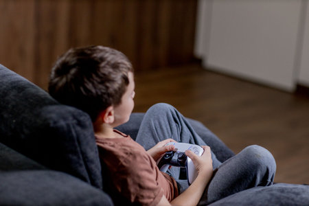 Boy is sitting on sofa at home and playing video games with his joypadの写真素材