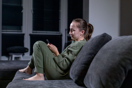 A woman is comfortably seated on a soft couch as she gazes intently at her smartphone, surrounded by the cozy ambiance of her living spaceの写真素材