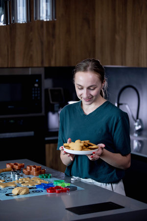 A joyful young girl is happily holding a plate full of freshly baked cookies in a modern kitchen...