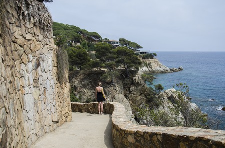 Panorama of Beautiful rocks road in Lloret de Mar and attractive young woman walking in black dress and with a yellow bag looking on a sea. Girl stands against rolling sea and rocks in Spain.の写真素材