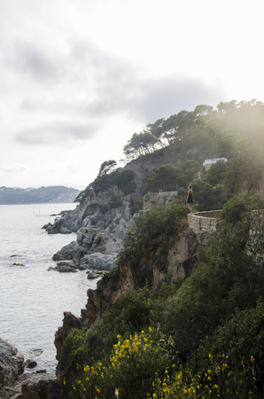 Attractive young woman in black dress and with a yellow little bag looking on a sea in sunset. Slim beautiful girl dress stands against rolling sea and beautiful rocks in Spain, Lloret de Mar.の写真素材