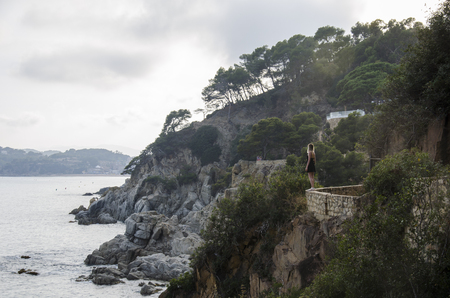 Attractive young woman in black dress and with a yellow little bag looking on a sea in sunset. Slim beautiful girl dress stands against rolling sea and beautiful rocks in Spain, Lloret de Mar.の写真素材