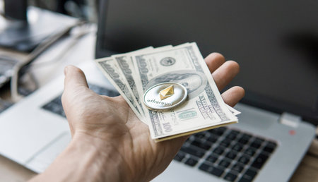 Close up man hand holding Ethereum coin with a us dollar bill against a laptop on the back. Ethereum...