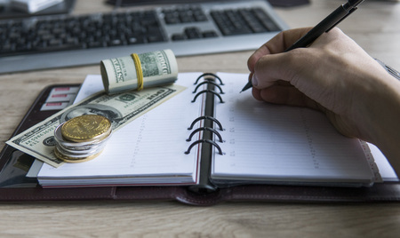 Bitcoin crypto currency on top dollar biils on notepad with mans hand with pen. Profit from mining crypto currencies. Miner with dollars from trading. Record of money income in notebook from mining.の写真素材