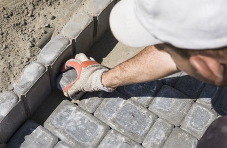 Hands of a builder in his orange gloved hands fitting laying new exterior paving stones carefully placing one in position on a leveled and raked soil base. Sand foundation. Building.の写真素材