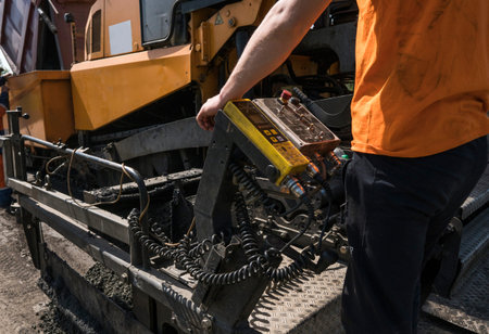 Worker operating asphalt paver machine during road construction and repairing works. A paver...