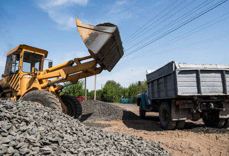 Yellow loader delivering stone gravel into truck during road construction works. The stones for the...